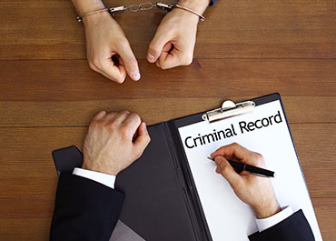 Overhead shot of hands in handcuffs on a wooden table, with a lawyer writing 'Criminal Record' on a document