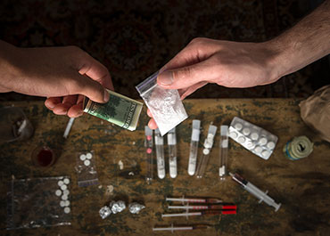 Hands exchanging money for a bag of white powder, with drug paraphernalia on a table, representing drug-related transactions