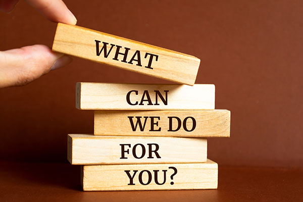 Hand holding a wooden block with 'WHAT' on top of stacked blocks spelling 'CAN WE DO FOR YOU?
