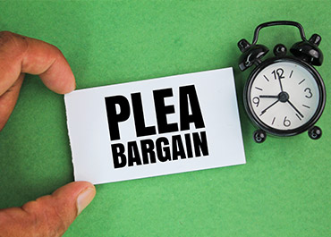 A hand holds a PLEA BARGAIN sign next to an alarm clock on a green background.