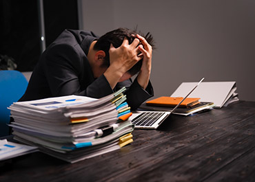 A man looking distressed with his hands on his head, surrounded by stacks of papers and a laptop.
